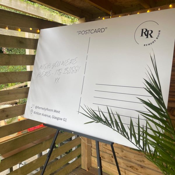 Giant postcard prop, with 'Wish you were here, it's bliss' written on the left, and the Remedy Room West logo on the righthand corner.